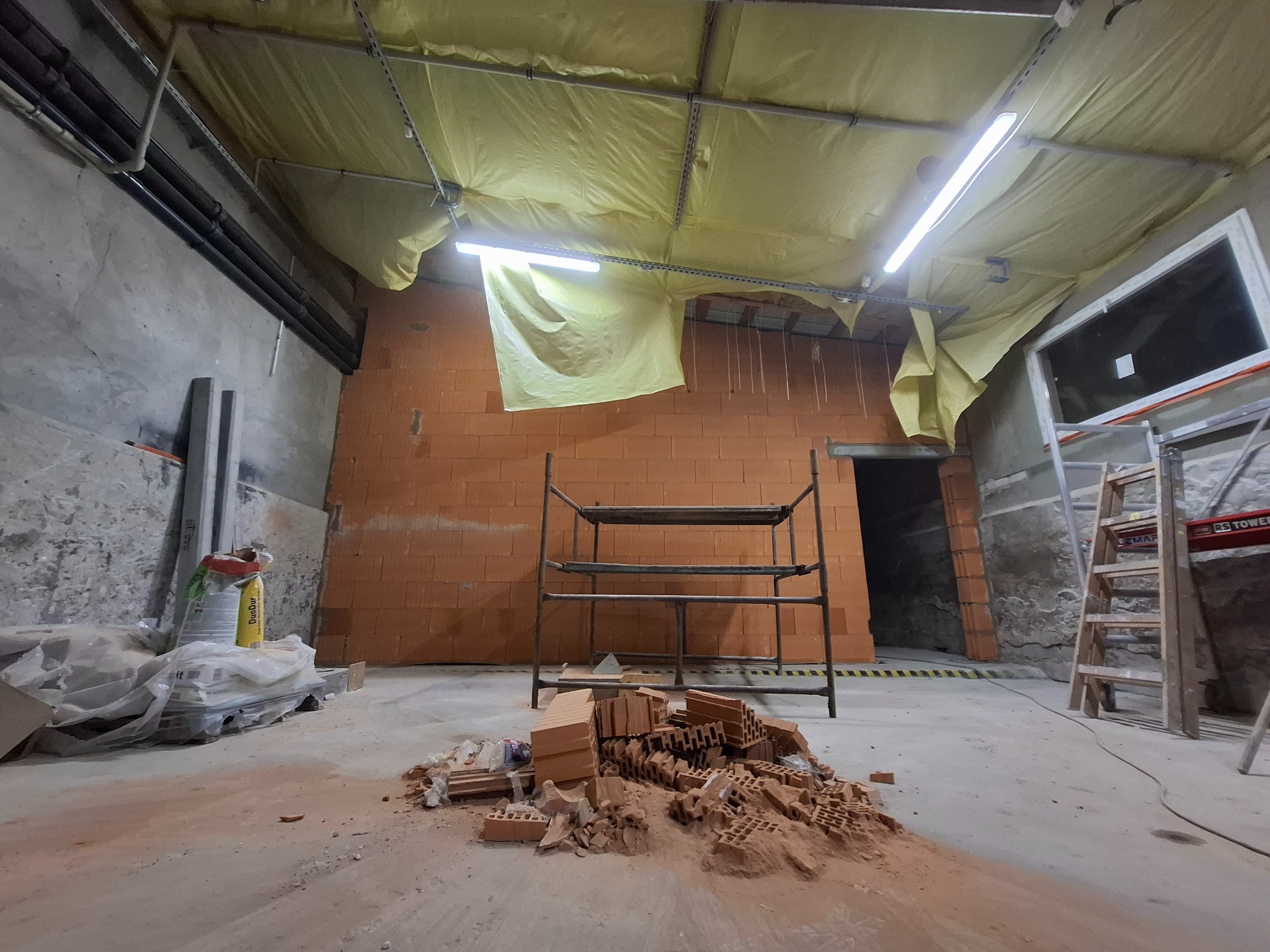 Wnętrze w trakcie budowy: ściana z czerwonej cegły, rozsypane cegły na podłodze, rusztowanie, drabina, okno i instalacje pod sufitem zabezpieczonym folią.