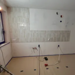 Ściana w trakcie układania beżowych, podłużnych płytek, widoczne przygotowane instalacje elektryczne i hydrauliczne, zabezpieczona podłoga i okno, lampa budowlana.