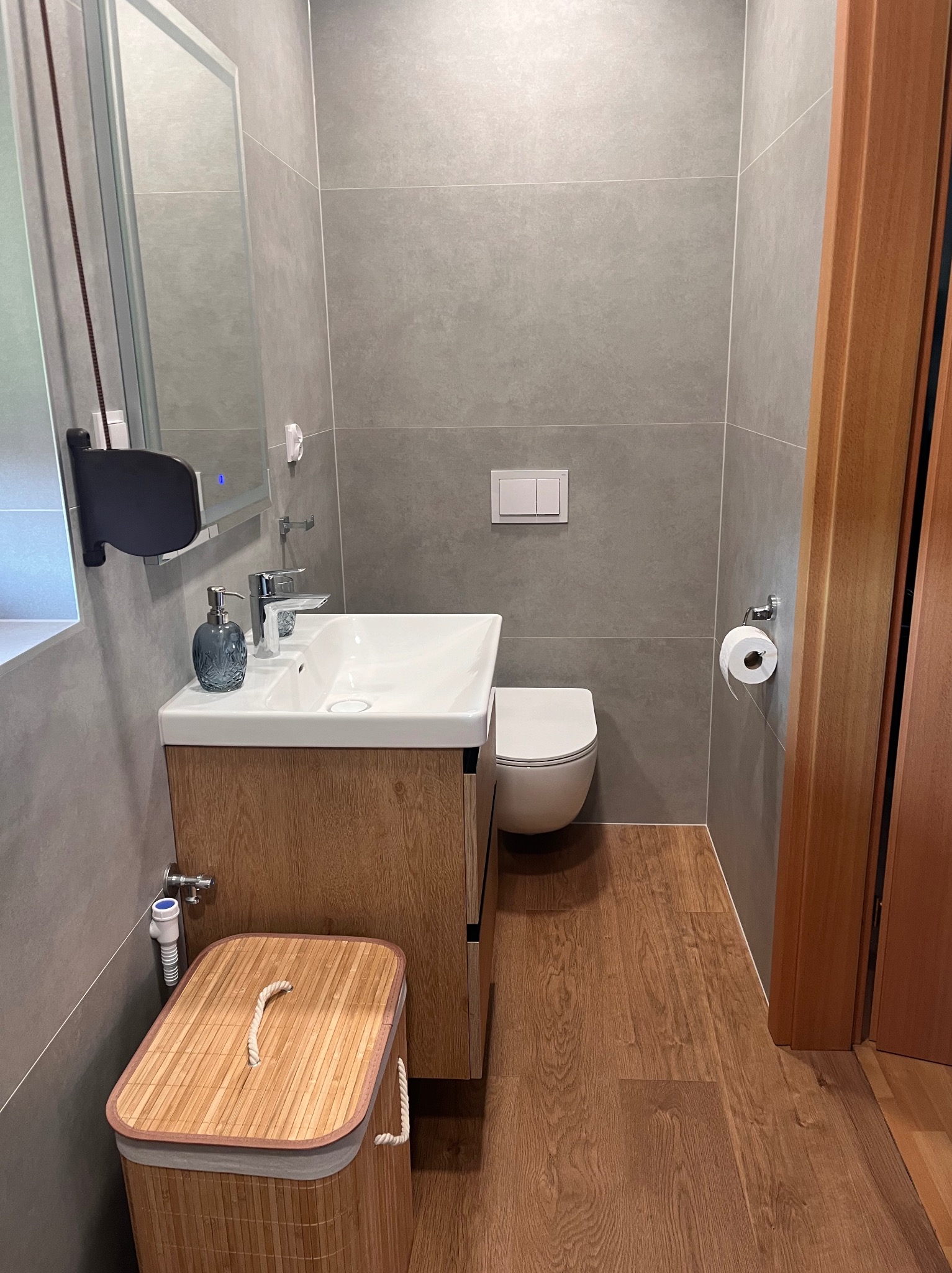 Nowoczesna łazienka z szarymi płytkami, drewnopodobną podłogą, białą umywalką z szafką z drewnianym wzorem, wiszącą toaletą i bambusowym koszem na pranie.