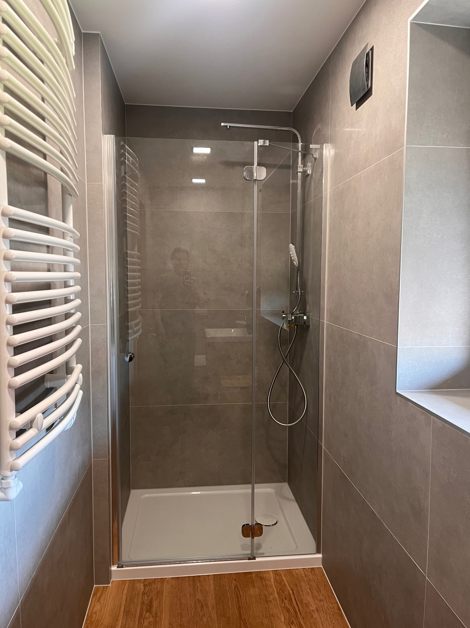 Nowoczesna kabina prysznicowa z przesuwnymi drzwiami, zamontowana w łazience z szarymi płytkami i drewnopodobną podłogą, widoczny grzejnik drabinkowy i zestaw prysznicowy.