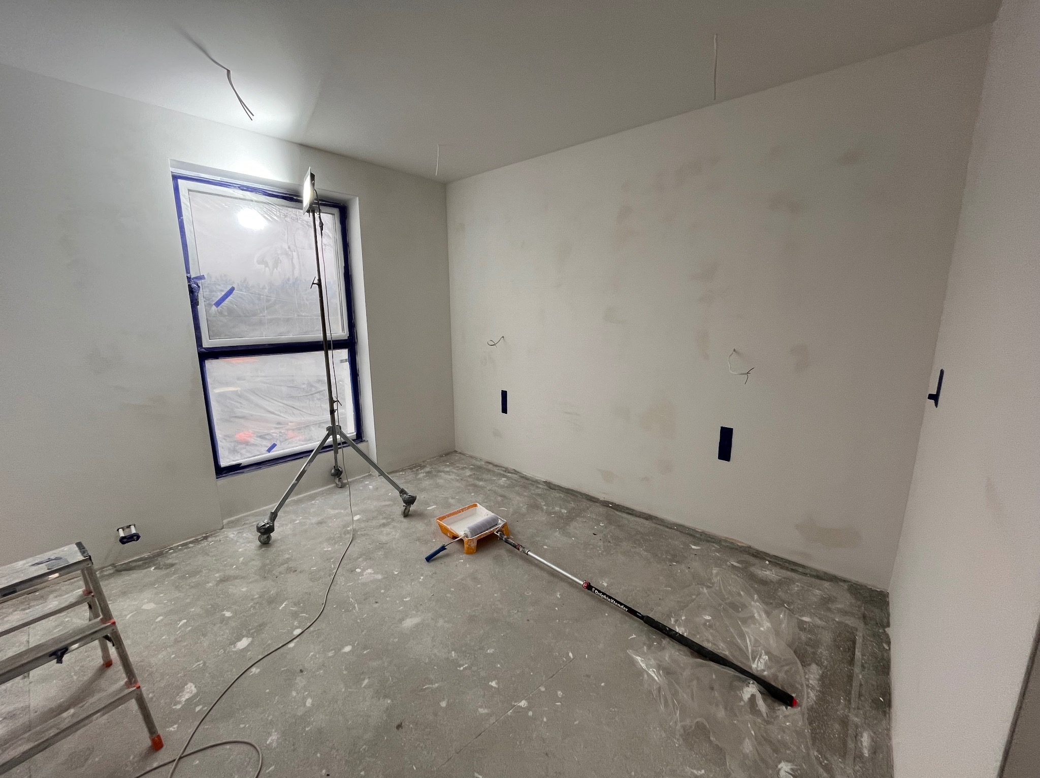 Pokój w Wiązowie w trakcie przygotowania do malowania: ściany po szpachlowaniu, zabezpieczone okno folią, widoczny wałek malarski z kuwetą, lampa oświetleniowa na statywie i drabina.