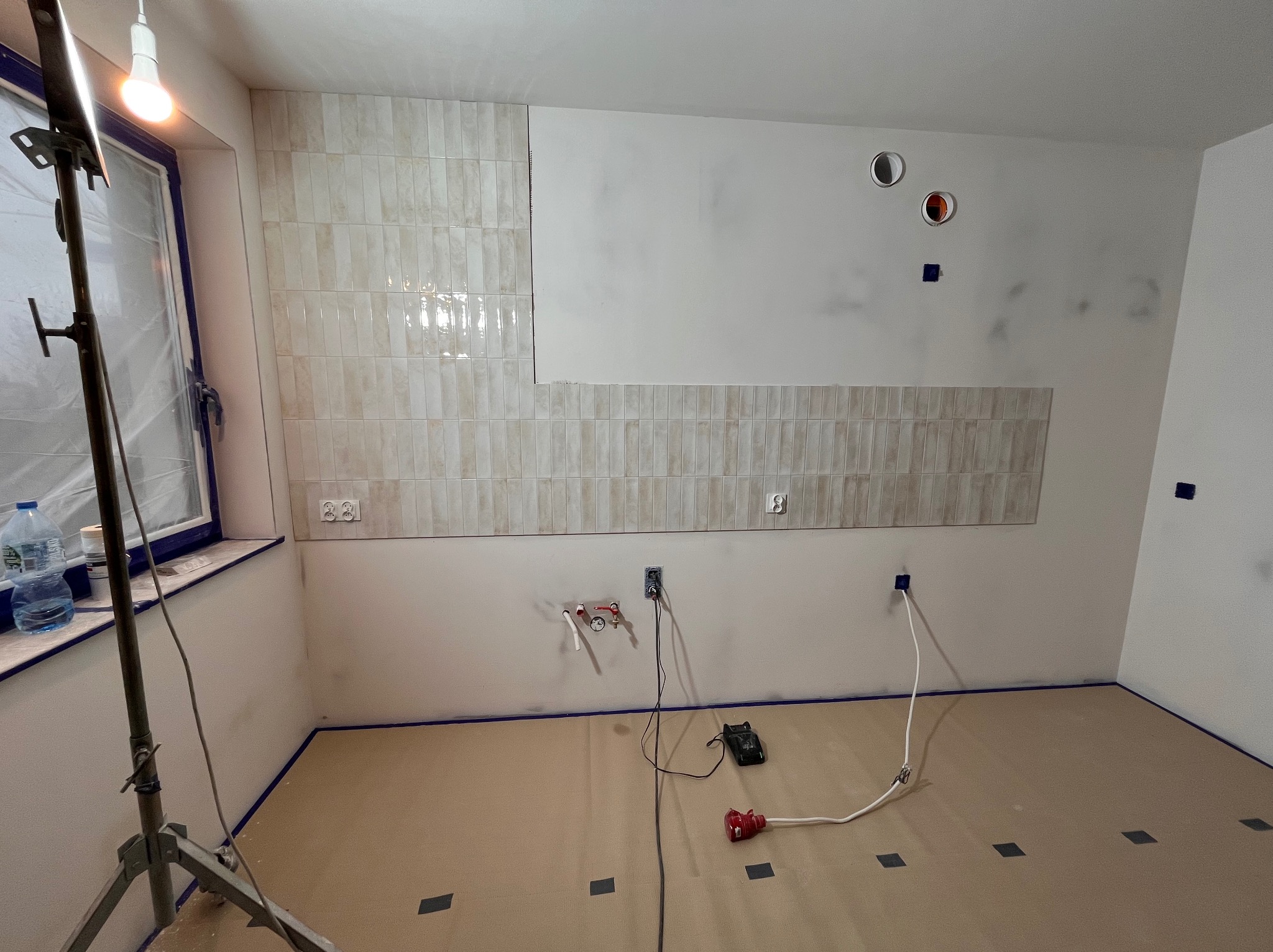 Ściana w trakcie układania beżowych, podłużnych płytek, widoczne przygotowane instalacje elektryczne i hydrauliczne, zabezpieczona podłoga i okno, lampa budowlana.