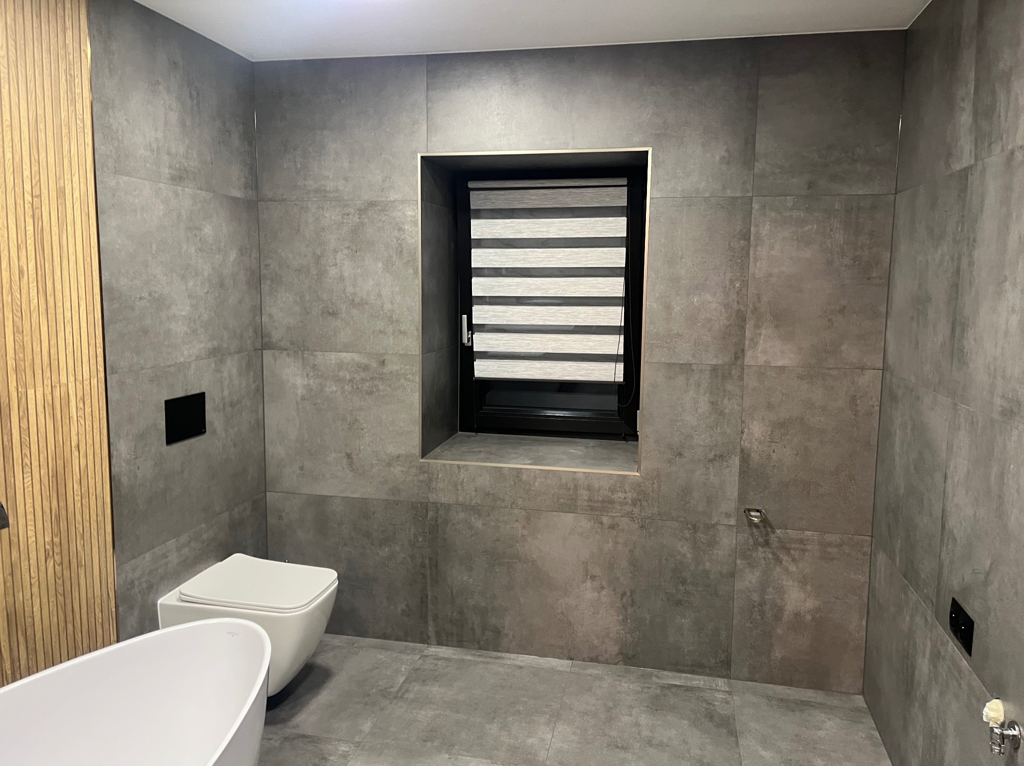 Nowoczesna łazienka z szarymi, wielkoformatowymi płytkami, minimalistyczną toaletą, wolnostojącą wanną i oknem z roletami dzień-noc.