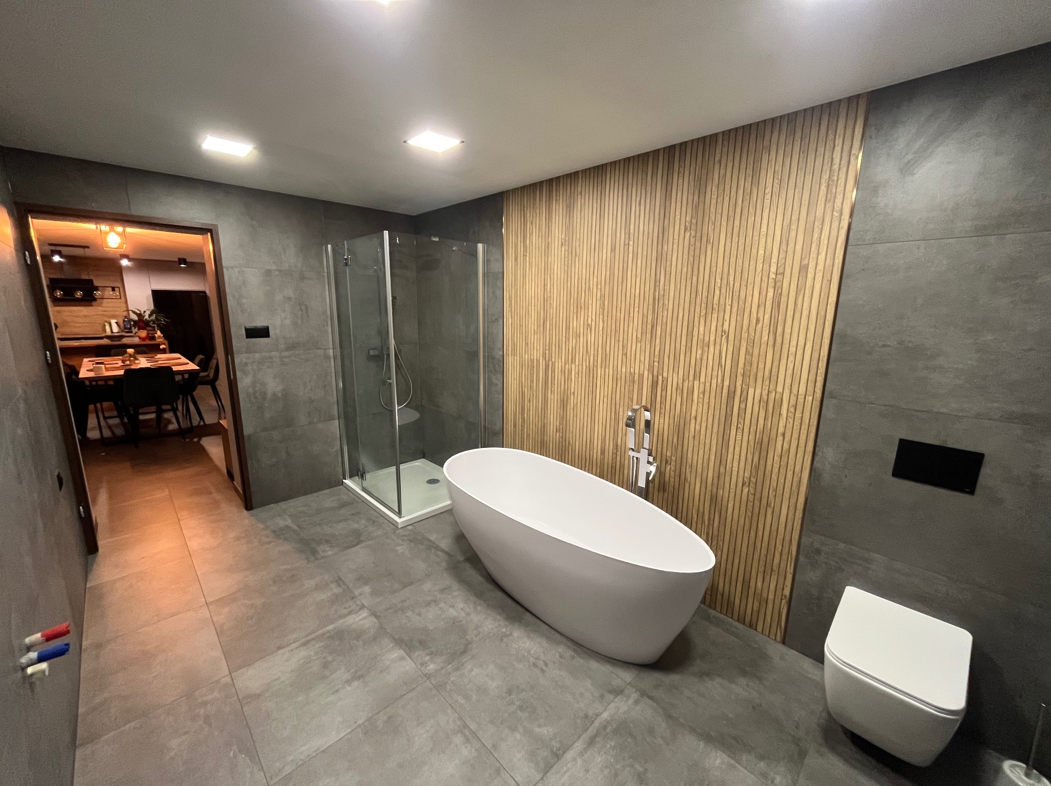 Nowoczesna łazienka z wolnostojącą wanną, kabiną prysznicową, podwieszaną toaletą i drewnianą ścianą dekoracyjną z listew, widok na kuchnię w tle.