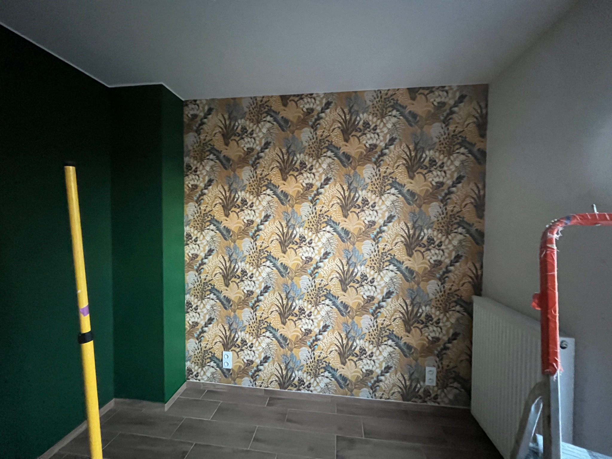 Pokój po remoncie: ściana z wzorzystą tapetą w odcieniach brązu, żółci i szarości, sąsiadująca z ciemnozieloną ścianą, widoczny fragment grzejnika i drabiny malarskiej.