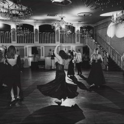 Mikołaj Musiałek Fotografia - Czarno-białe zdjęcie sali balowej podczas studniówki: tańczący ludzie, eleganckie suknie, żyrandole, schody i balkon. Dynamiczne ujęcie ruchu i atmosfery.