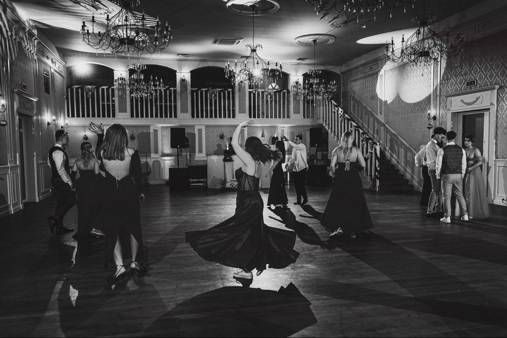 Czarno-białe zdjęcie sali balowej podczas studniówki: tańczący ludzie, eleganckie suknie, żyrandole, schody i balkon. Dynamiczne ujęcie ruchu i atmosfery.