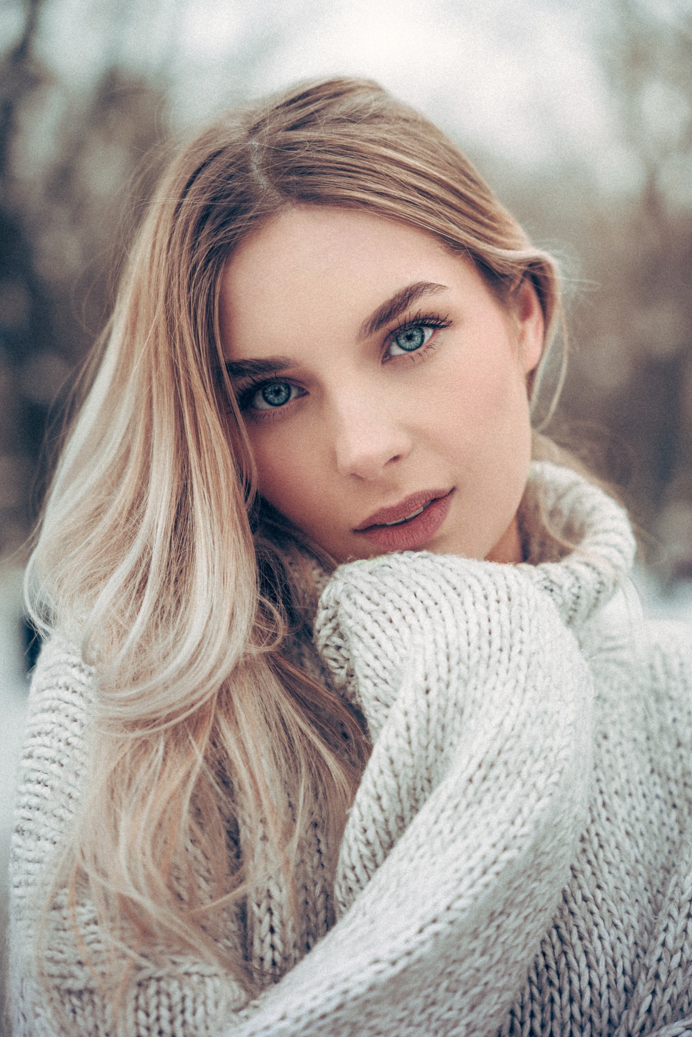 Portret młodej blondynki o niebieskich oczach w grubym, kremowym swetrze na tle rozmytych drzew w zimowej scenerii, delikatne światło, subtelny makijaż.