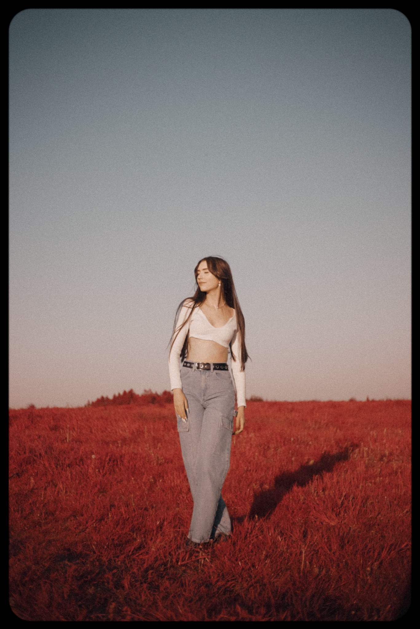 Portret kobiety w plenerze na tle czerwonej trawy. Ubrana w biały top i jeansy, z długimi brązowymi włosami, odwraca wzrok w lewo. Delikatne światło, naturalny wygląd.