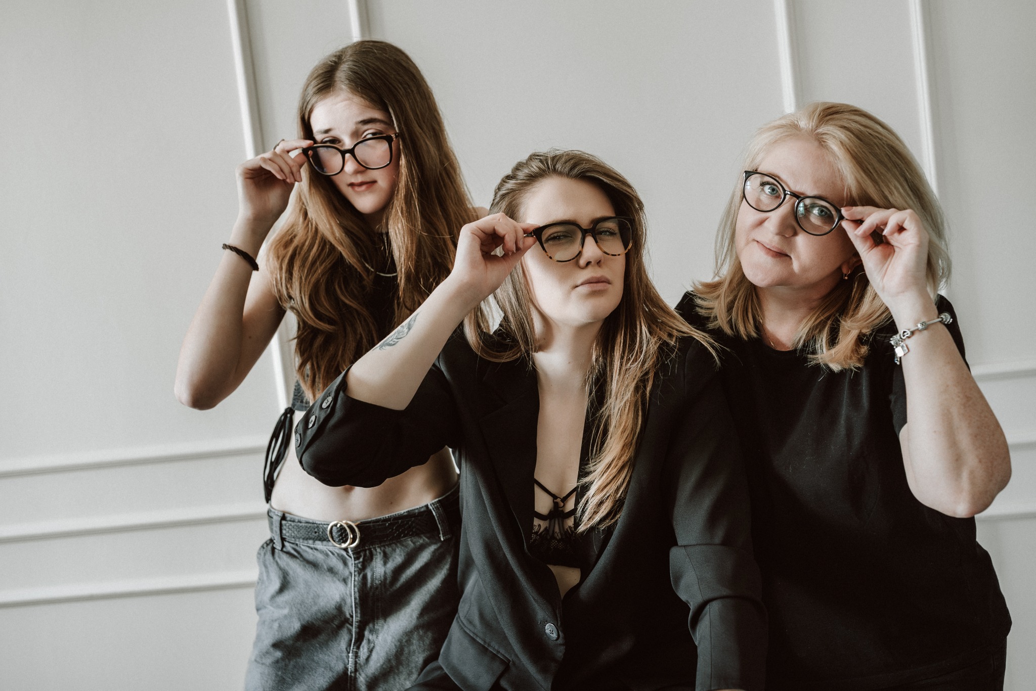 Portret trzech kobiet w okularach na tle białej ściany. Różne style ubioru i wyraz twarzy. Rodzinna sesja w neutralnym oświetleniu.