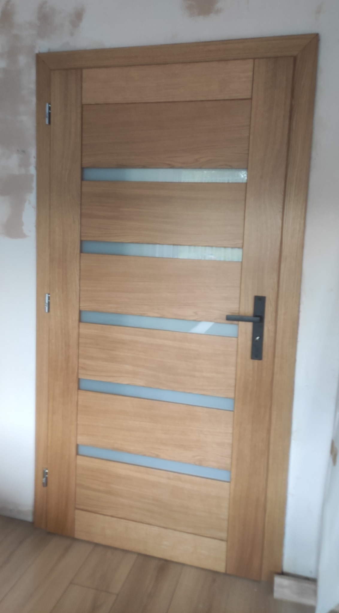 Jasne, drewniane drzwi wewnętrzne z poziomymi, matowymi przeszkleniami i czarną klamką, widoczne na tle ściany w trakcie remontu.
