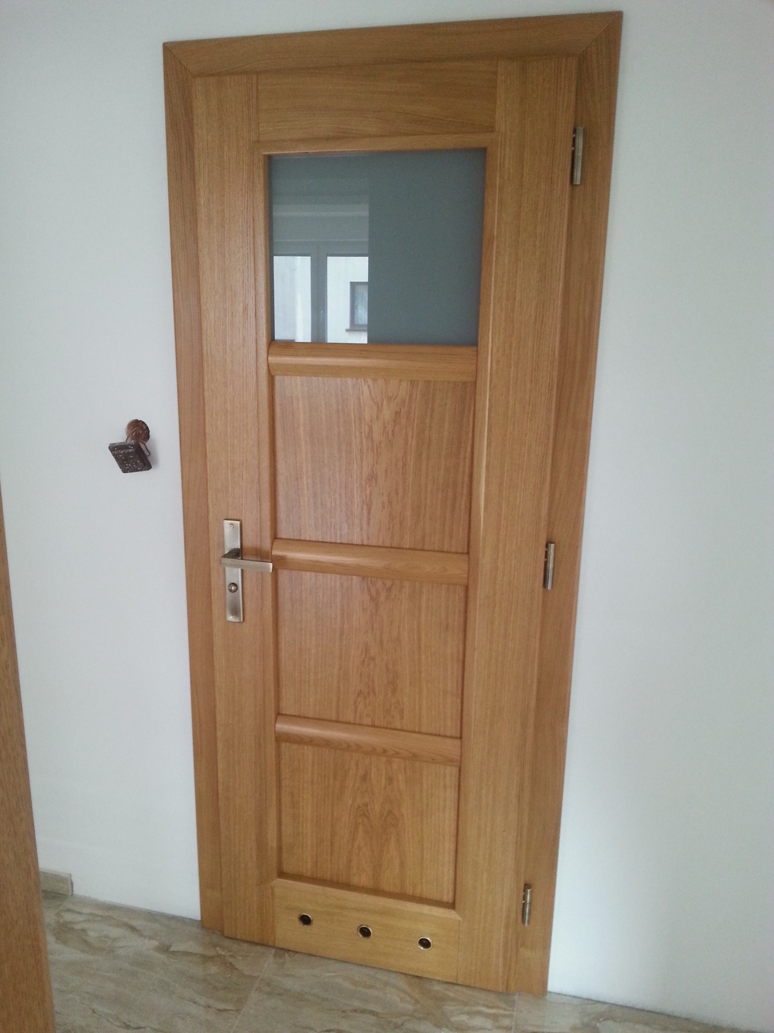 Jasne, drewniane drzwi wewnętrzne z prostokątnym przeszkleniem u góry, srebrna klamka i widoczne zawiasy, zamontowane w białej ścianie nad marmurową podłogą.