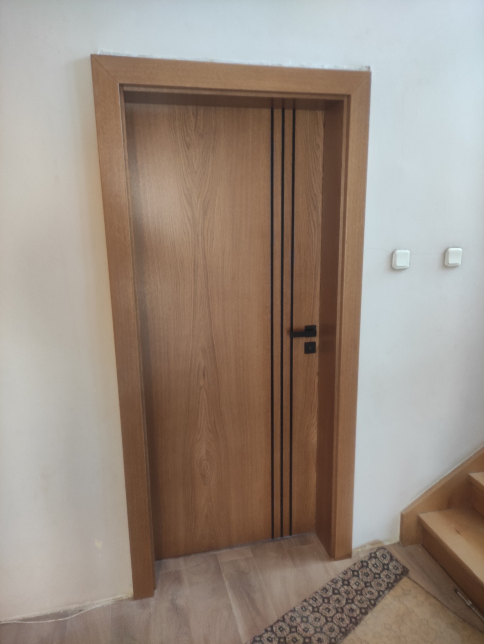 Nowoczesne drzwi wewnętrzne z jasnego drewna z trzema pionowymi, czarnymi paskami, czarną klamką na tle białej ściany i fragmentu schodów.