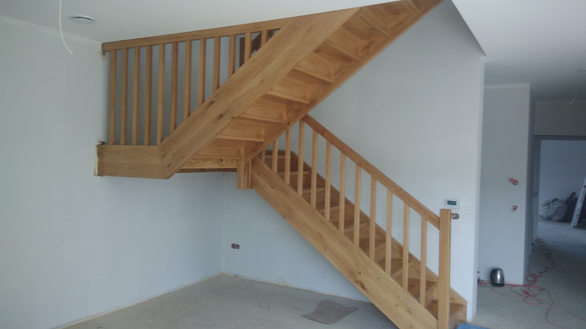 Jasne drewniane schody z balustradą w surowym stanie, widoczne niedokończone wykończenia ścian i podłóg, w tle widoczne kable i narzędzia.