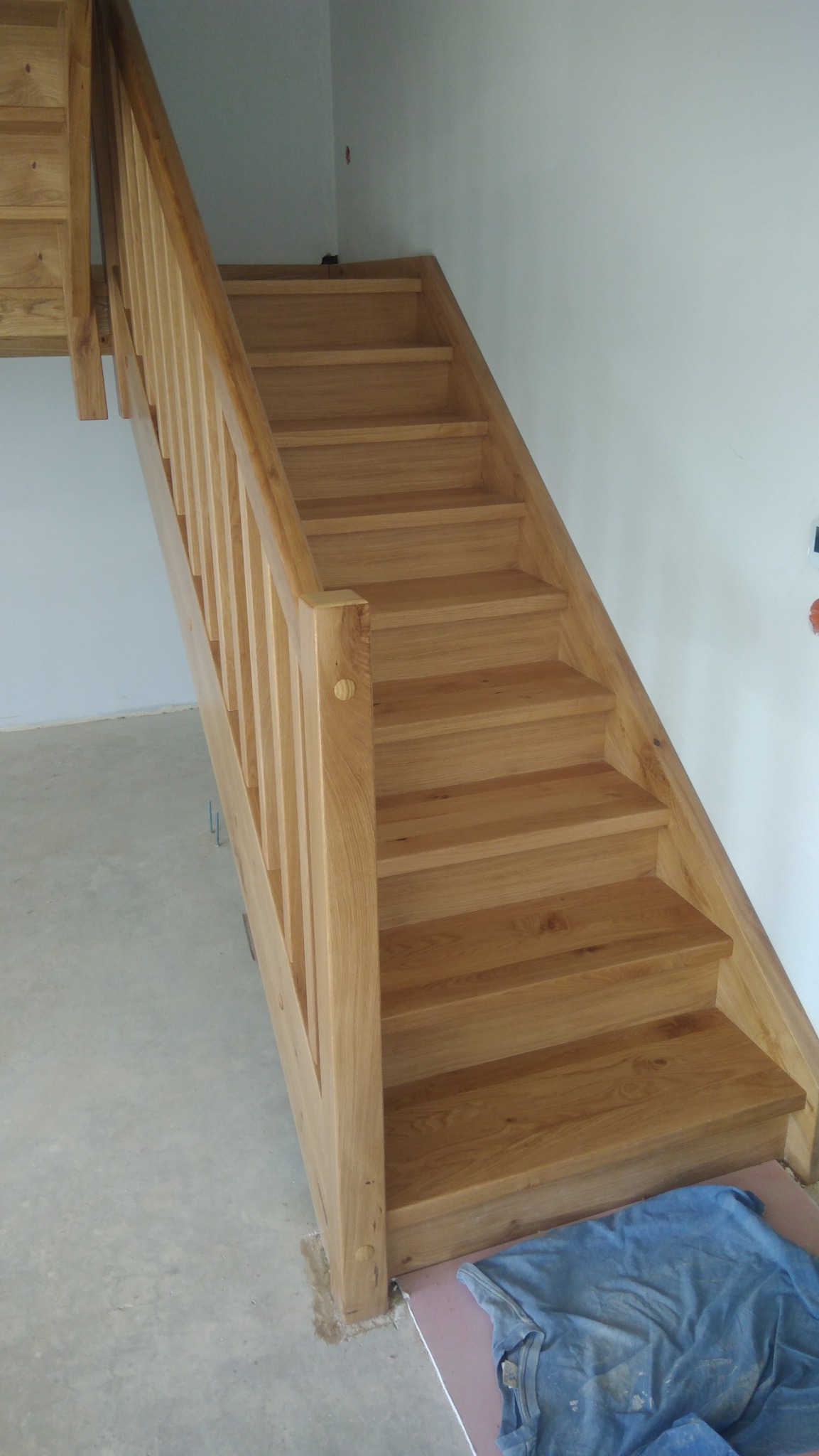 Jasne, drewniane schody z balustradą w trakcie montażu, widoczna niebieska szmatka na podłodze i fragment surowej ściany.