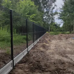 Długi fragment czarnego ogrodzenia panelowego z betonową podmurówką, widok z boku na tle drzew i ziemi po montażu.