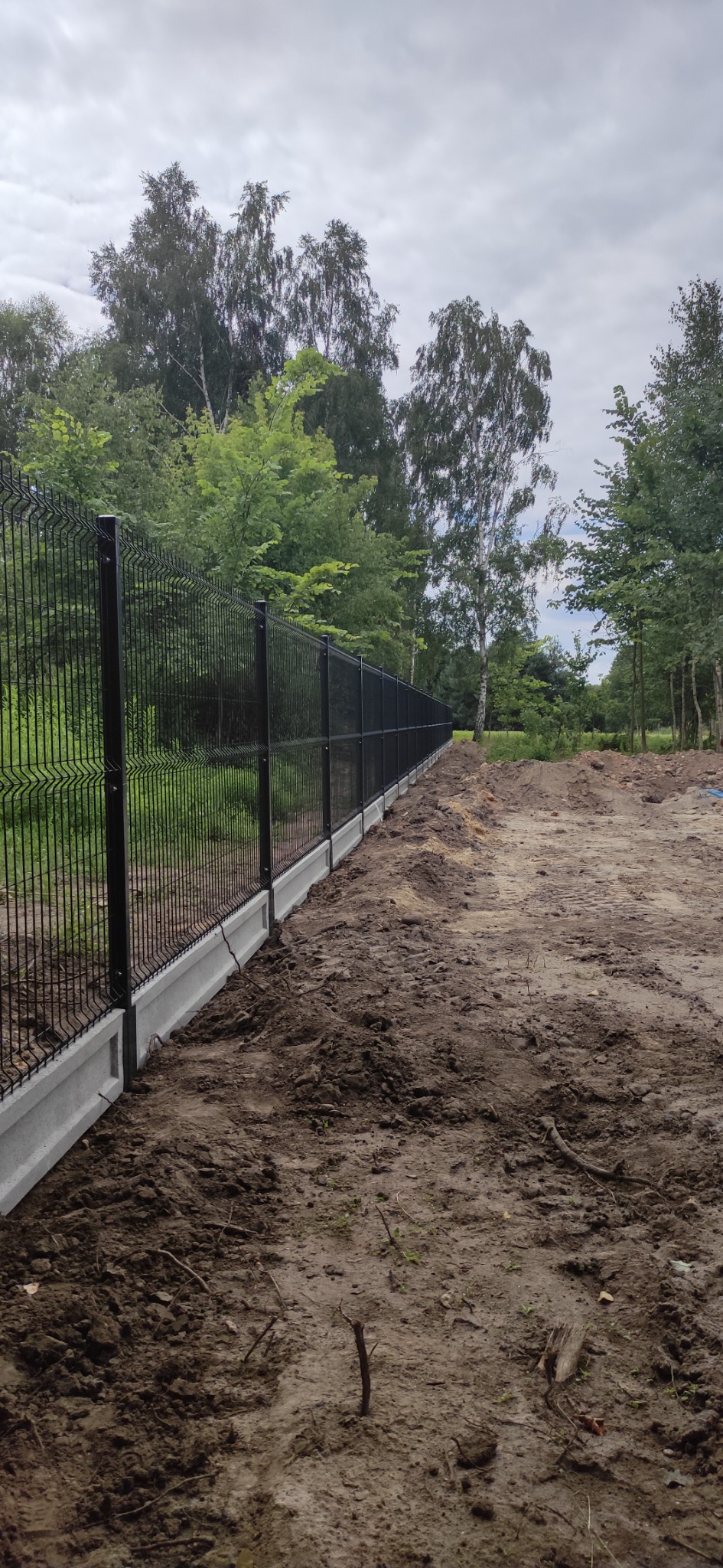 Długi fragment czarnego ogrodzenia panelowego z betonową podmurówką, widok z boku na tle drzew i ziemi po montażu.