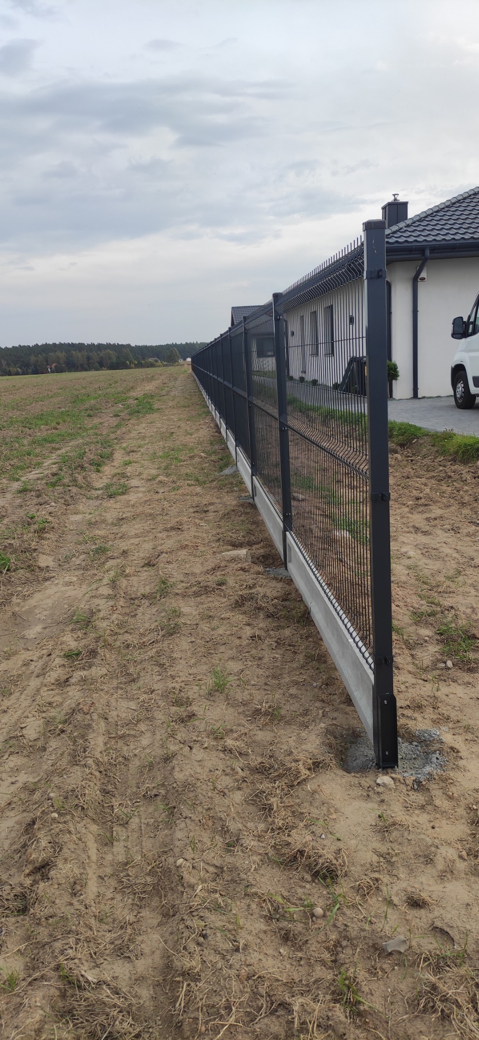 Długie, metalowe ogrodzenie panelowe z podmurówką betonową, oddzielające posesję od pola, widok z boku w pochmurny dzień.