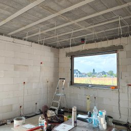 BarTech - Wnętrze w trakcie montażu instalacji elektrycznej: przewody zwisają z sufitu, widoczne puszki w ścianach z betonu komórkowego, okno z widokiem na domy.