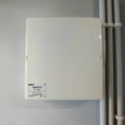 Zamknięta, biała skrzynka elektryczna Safel PERFECTA 16, zamontowana na szarej ścianie, obok białych rur instalacyjnych.