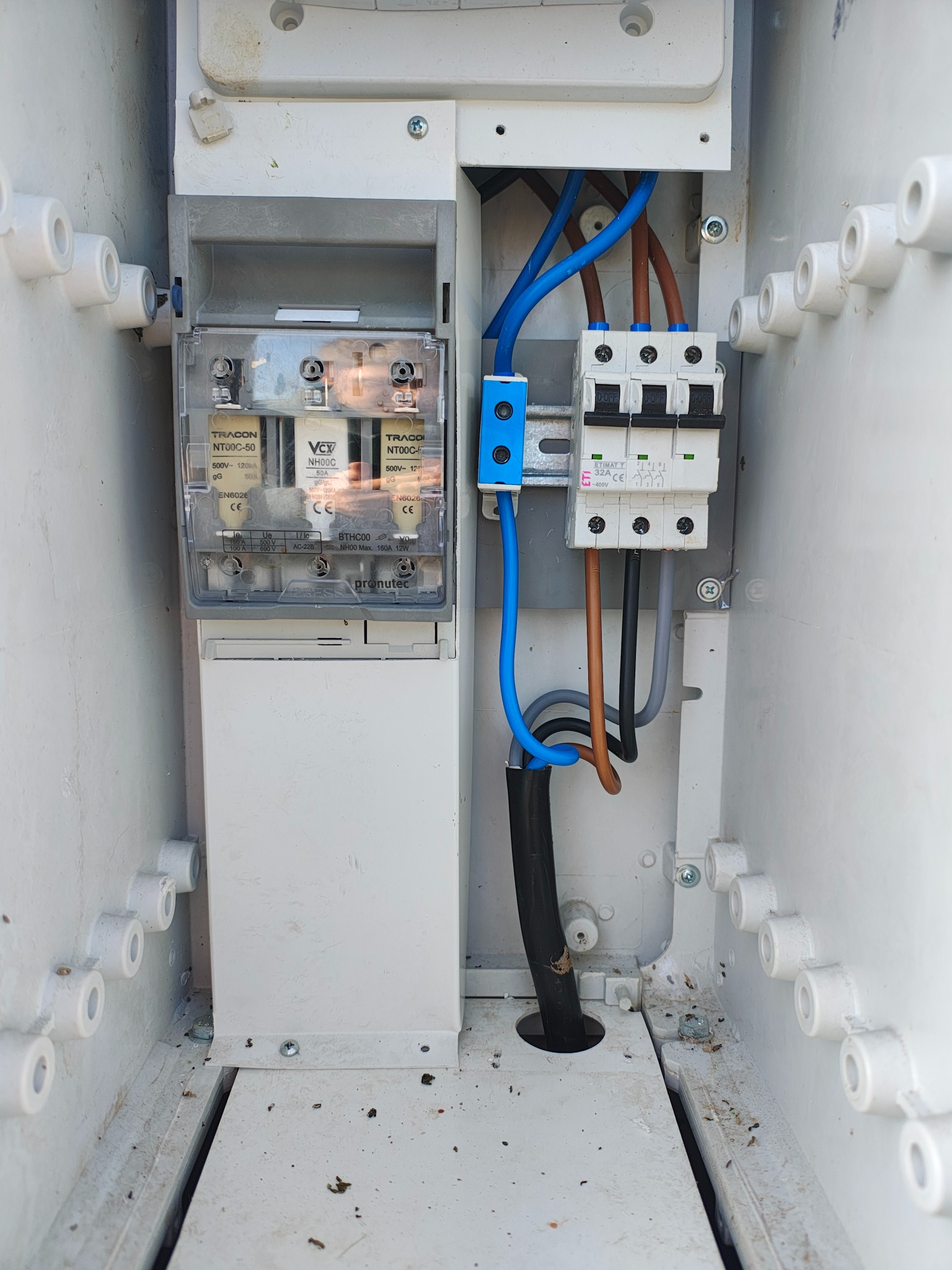 Wnętrze skrzynki elektrycznej z bezpiecznikami TRACON, wyłącznikiem ETIMAT i okablowaniem w kolorach niebieskim, brązowym i czarnym, zamontowane w białej obudowie.