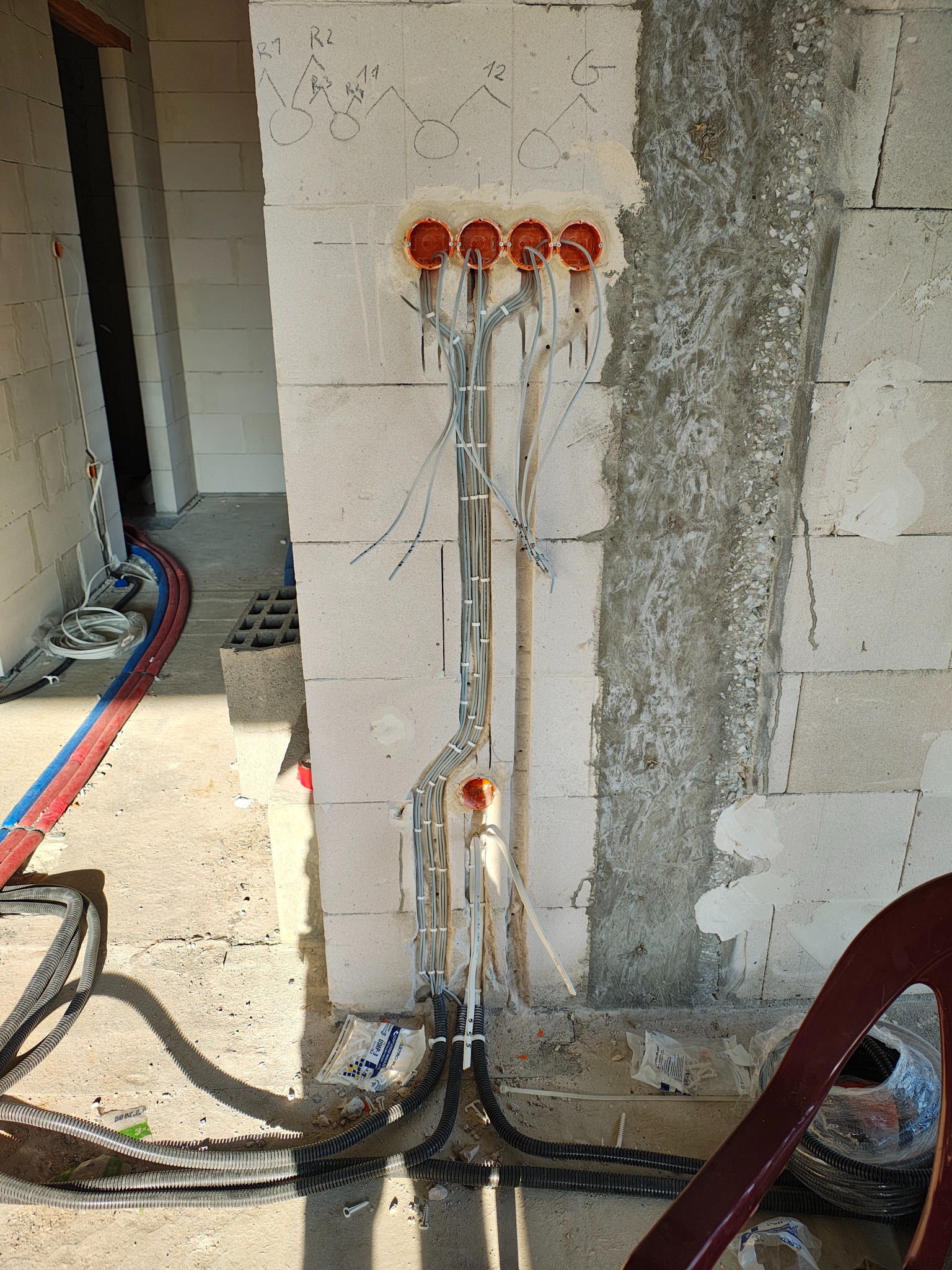 Wewnętrzna instalacja elektryczna w trakcie montażu: przewody w peszlach wychodzą z pomarańczowych puszek w ścianie z betonu komórkowego. Widoczne oznaczenia schematu.