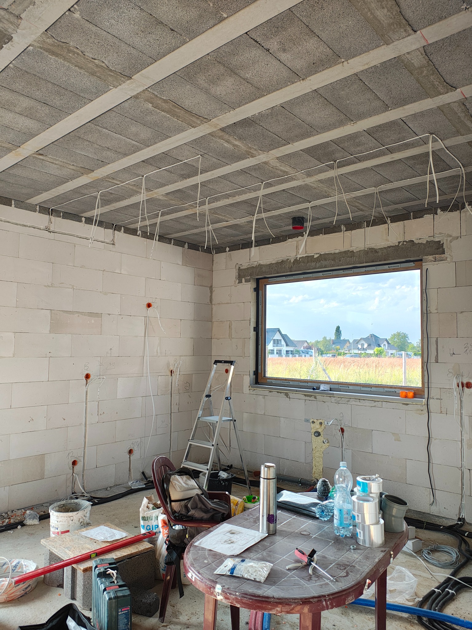 Wnętrze w trakcie montażu instalacji elektrycznej: przewody zwisają z sufitu, widoczne puszki w ścianach z betonu komórkowego, okno z widokiem na domy.