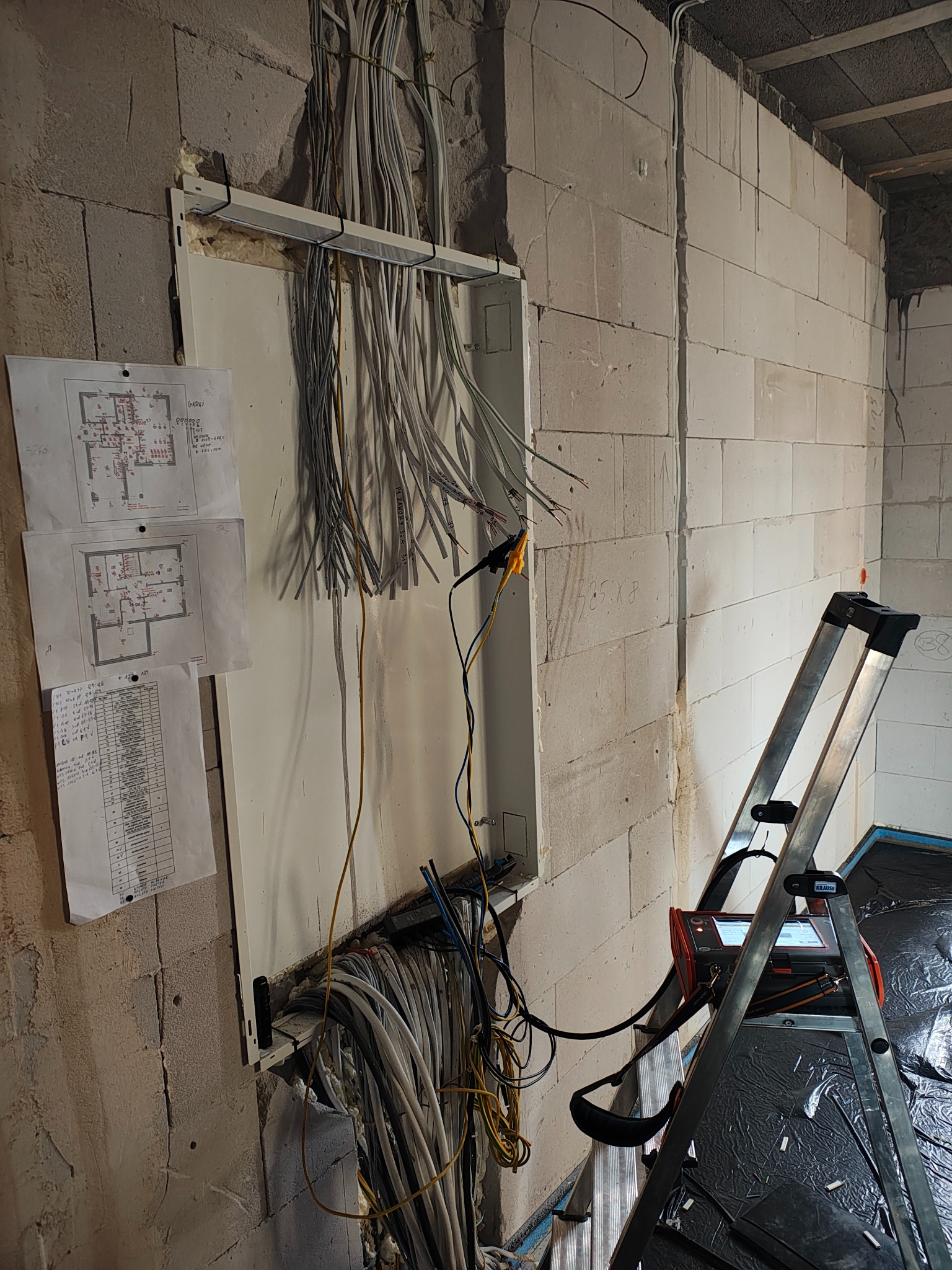 Rozdzielnia elektryczna w surowym stanie z wyprowadzonymi przewodami, schematy instalacji na ścianie, miernik na drabinie, blok komórkowy.