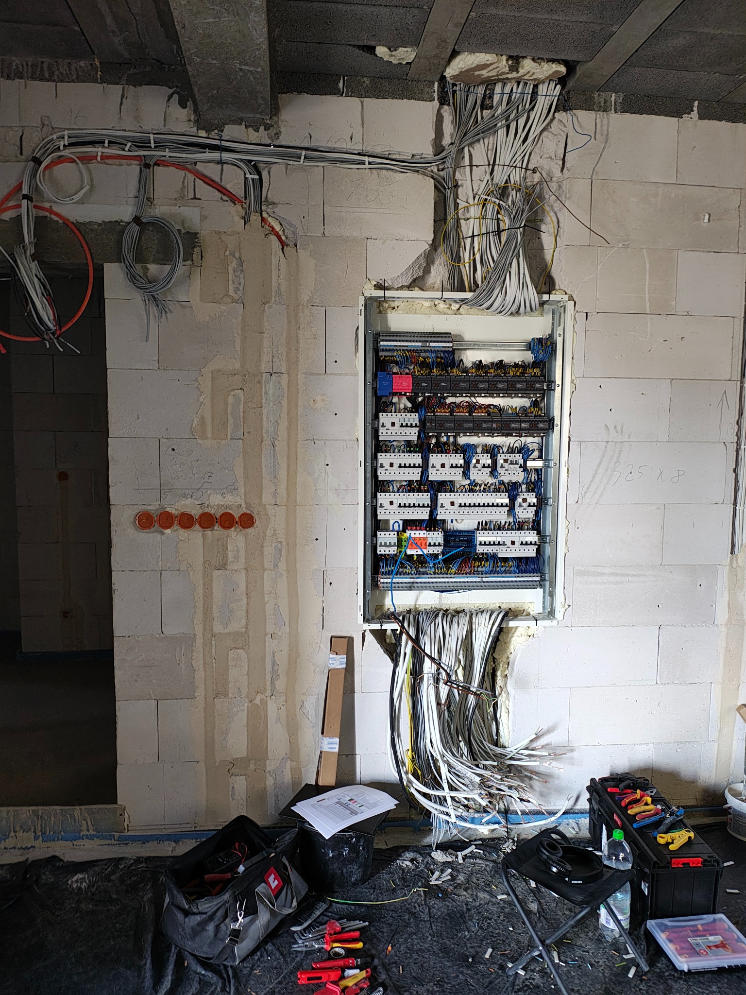 Rozdzielnia elektryczna w surowym stanie, z widocznymi kablami i przewodami w ścianie z betonu komórkowego. Na podłodze narzędzia i akcesoria.