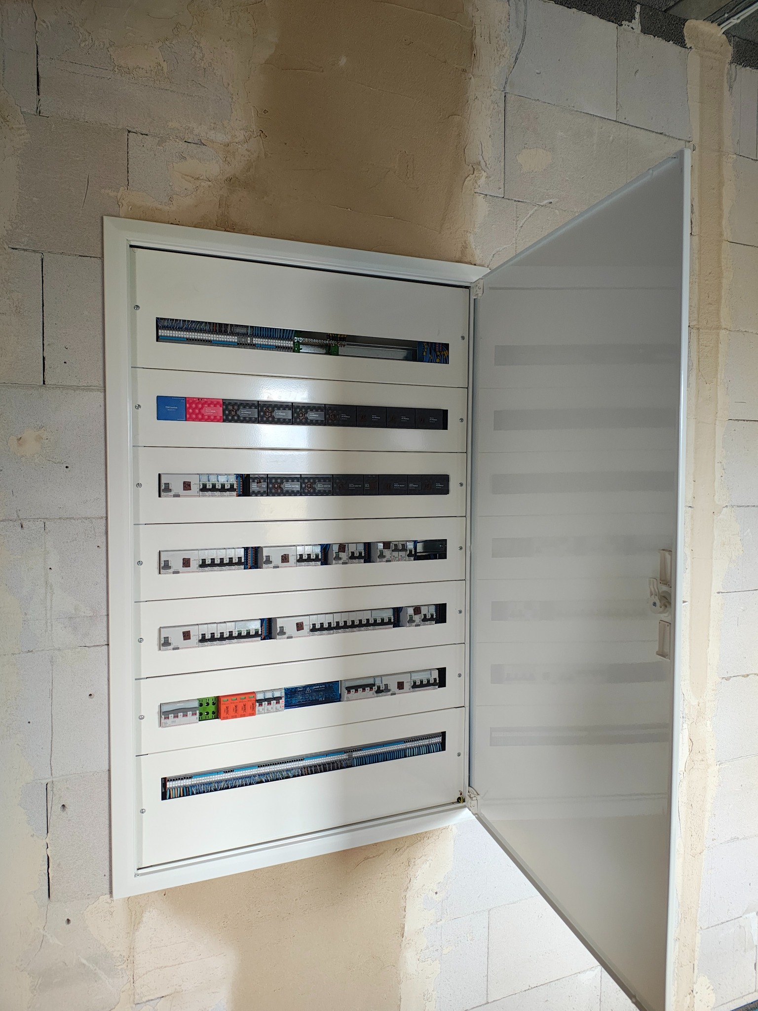 Otwarta, biała skrzynka rozdzielcza z bezpiecznikami na surowej ścianie z betonu komórkowego. Widoczne przewody i zabezpieczenia. Instalacja elektryczna w trakcie montażu.