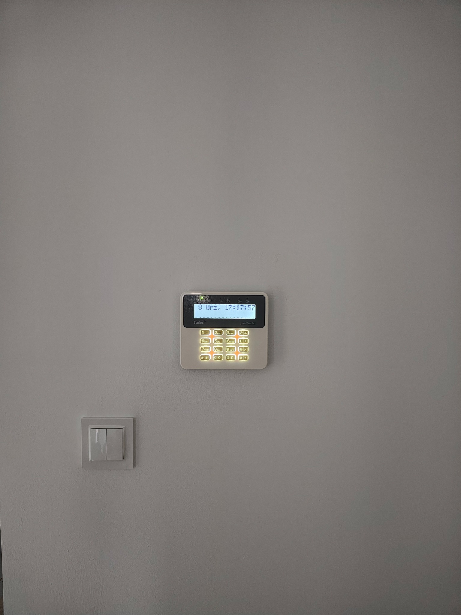 Panel alarmowy z podświetlanymi przyciskami zamontowany na ścianie obok włącznika światła. Widoczny ekran z datą i godziną. Minimalistyczny design.