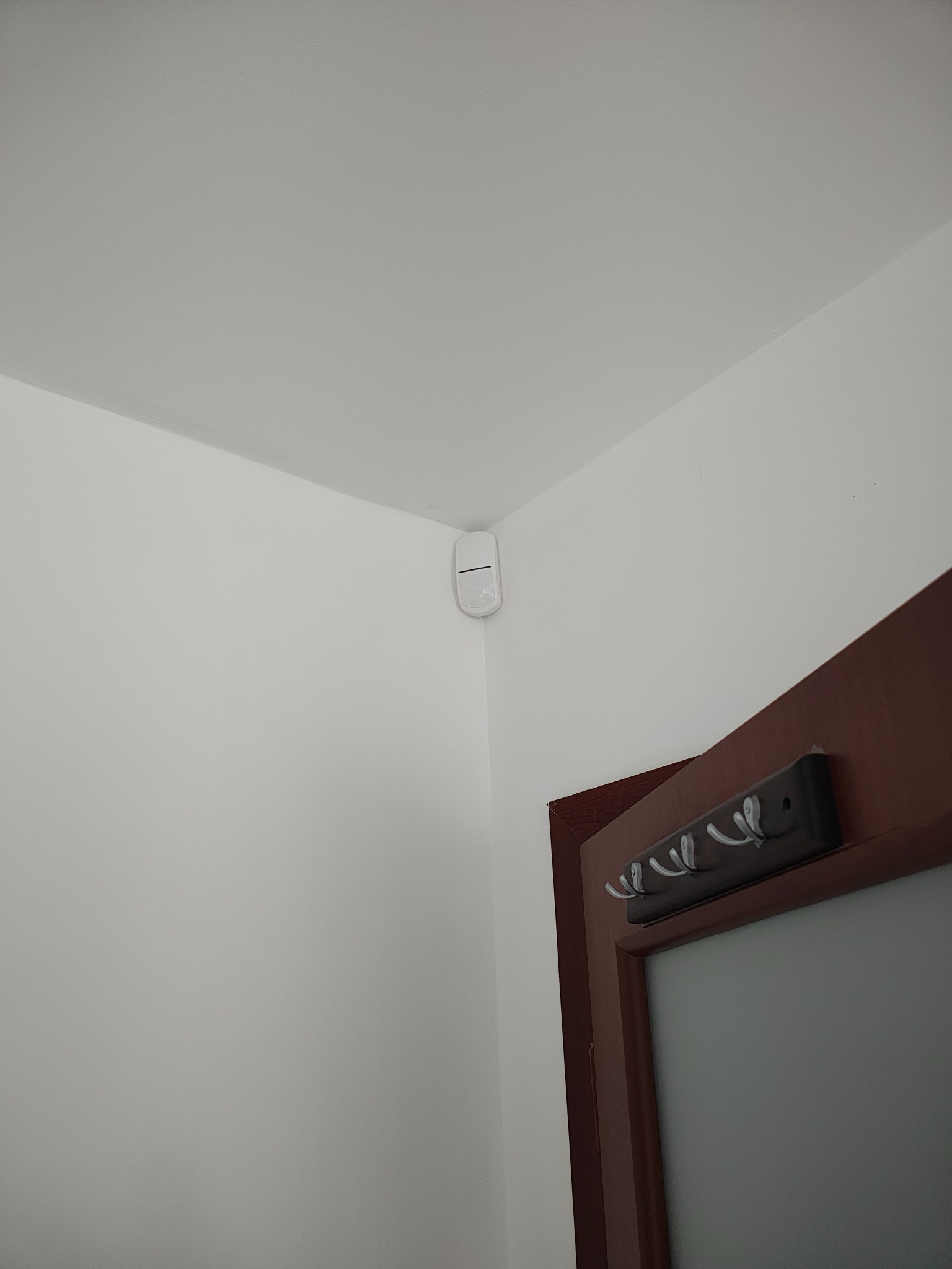 Biały czujnik ruchu zamontowany w rogu pokoju, obok framugi drzwi z wieszakami. Element systemu inteligentnego domu.