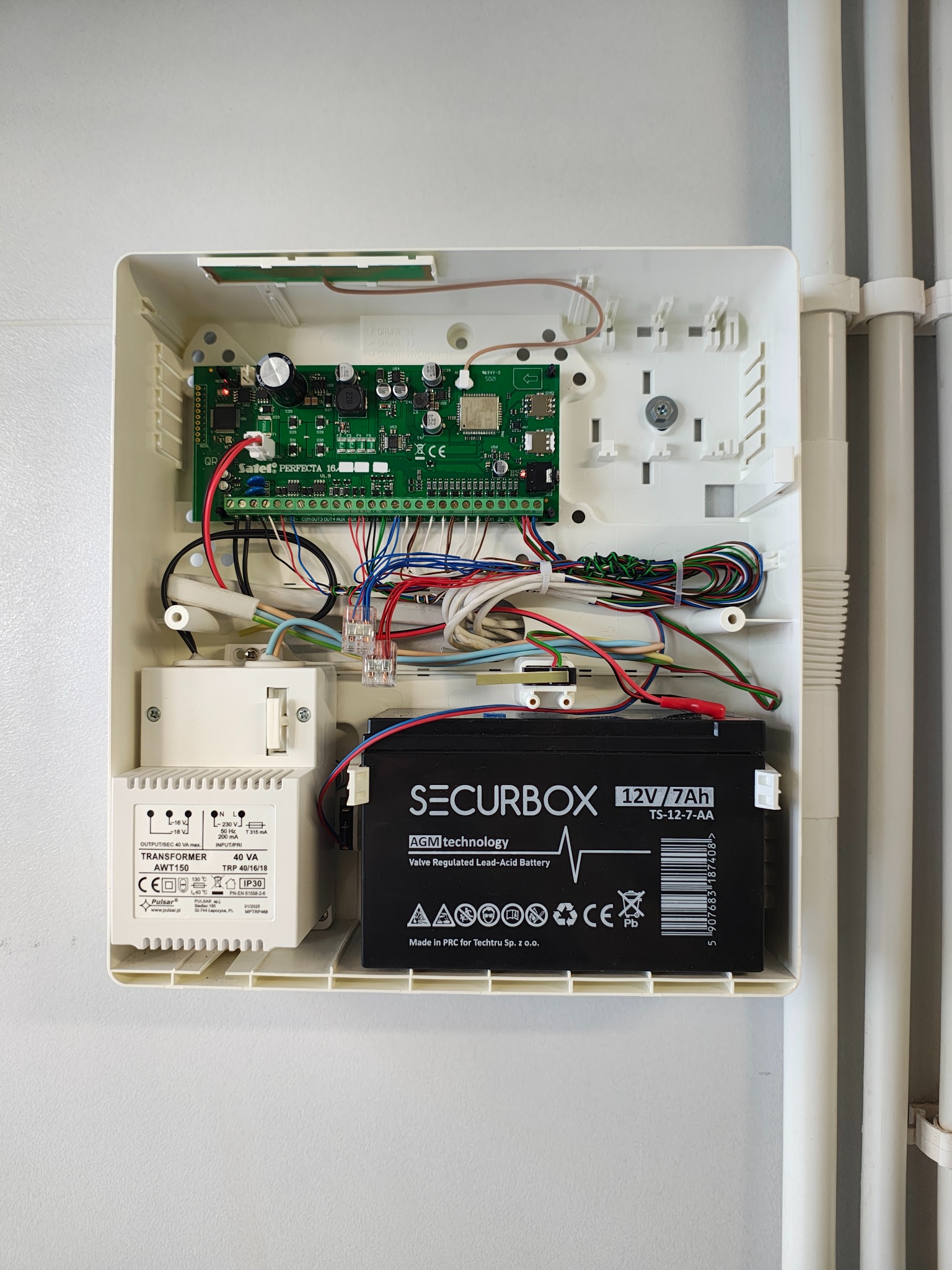 Otwarta skrzynka centrali alarmowej z akumulatorem, transformatorem i płytką drukowaną. Widoczne kable i przewody. Element instalacji zabezpieczeń.