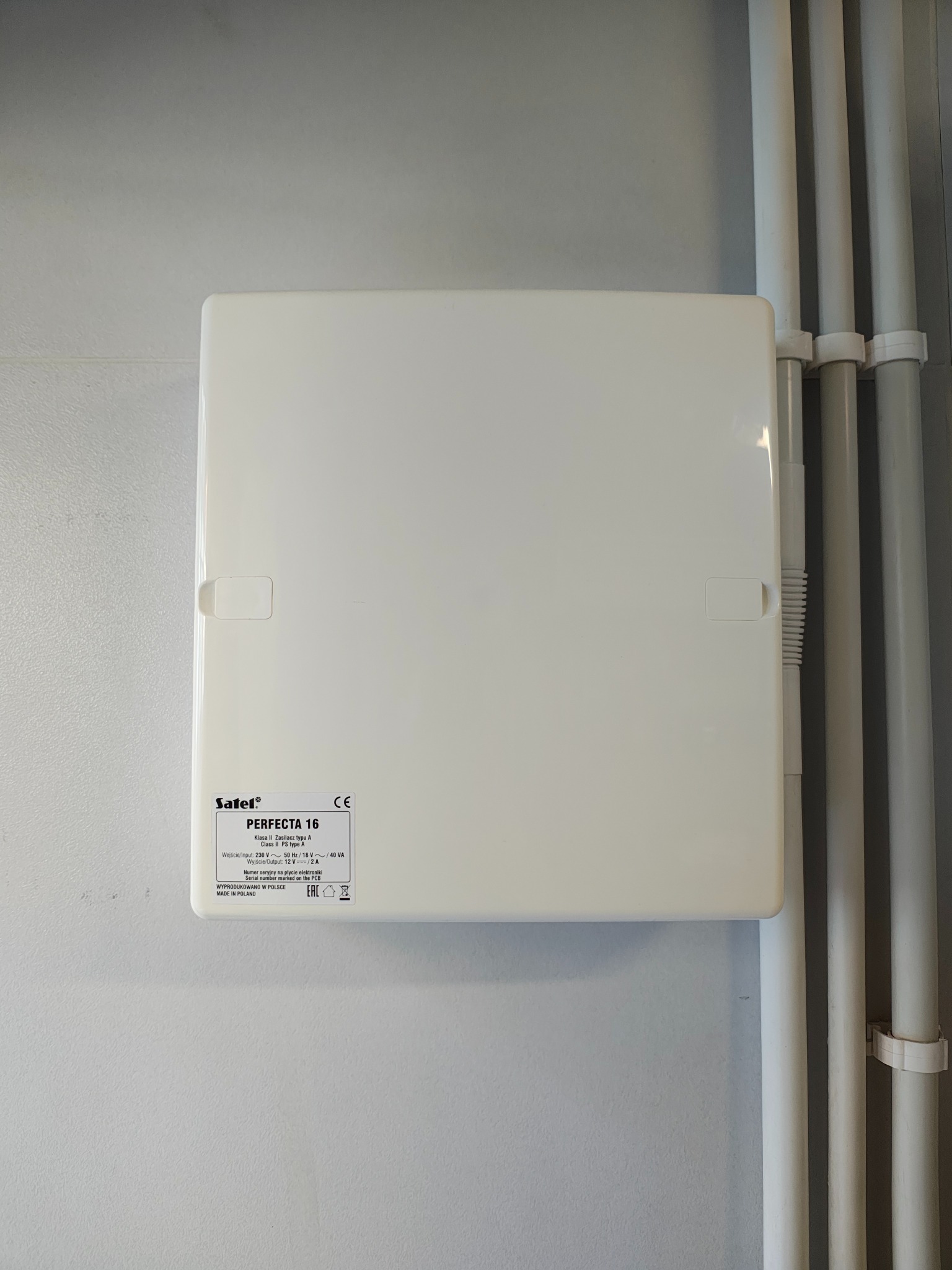 Zamknięta, biała skrzynka elektryczna Safel PERFECTA 16, zamontowana na szarej ścianie, obok białych rur instalacyjnych.