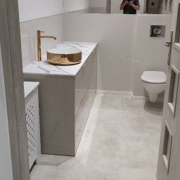 KAM-BUD Wykończenia wnętrz - Wykończona łazienka w odcieniach bieli i szarości z elementami złota: umywalka nablatowa w kolorze złotym, armatura w kolorze złotym, duże lustro, toaleta, płytki podłogowe w odcieniach szarości...