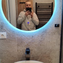 KAM-BUD Wykończenia wnętrz - Okrągłe lustro z niebieskim podświetleniem LED w wyremontowanej łazience, widoczna umywalka nablatowa, bateria w kolorze czarnym oraz fragment grzejnika drabinkowego na ścianie z płytek imitujących...