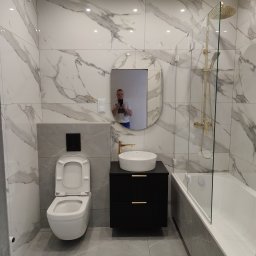 KAM-BUD Wykończenia wnętrz - Nowoczesna łazienka z marmurowymi płytkami, owalnym lustrem odbijającym fotografa, wiszącą toaletą, czarną szafką z umywalką i złotą armaturą oraz kabiną prysznicową ze złotym zestawem natryskowym.