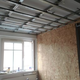 KAM-BUD Wykończenia wnętrz - Wnętrze pokoju w trakcie prac wykończeniowych: metalowy stelaż sufitu podwieszanego zamontowany, ściana wyłożona płytą OSB, okno z widocznymi przedmiotami na parapecie.