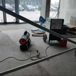 KAM-BUD Wykończenia wnętrz - Wnętrze w trakcie prac wykończeniowych, widoczny nagrzewnica elektryczna, agregat malarski na kółkach, worki z materiałami budowlanymi oraz fragment rusztowania na pierwszym planie.