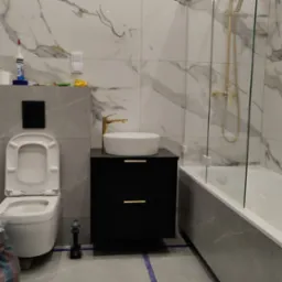 Nowoczesna łazienka w trakcie wykańczania: biała umywalka na czarnej szafce z złotą armaturą, toaleta z otwartą klapą, wanna za szklanym parawanem, marmurowe płytki na ścianach, widoczne narzędzia...