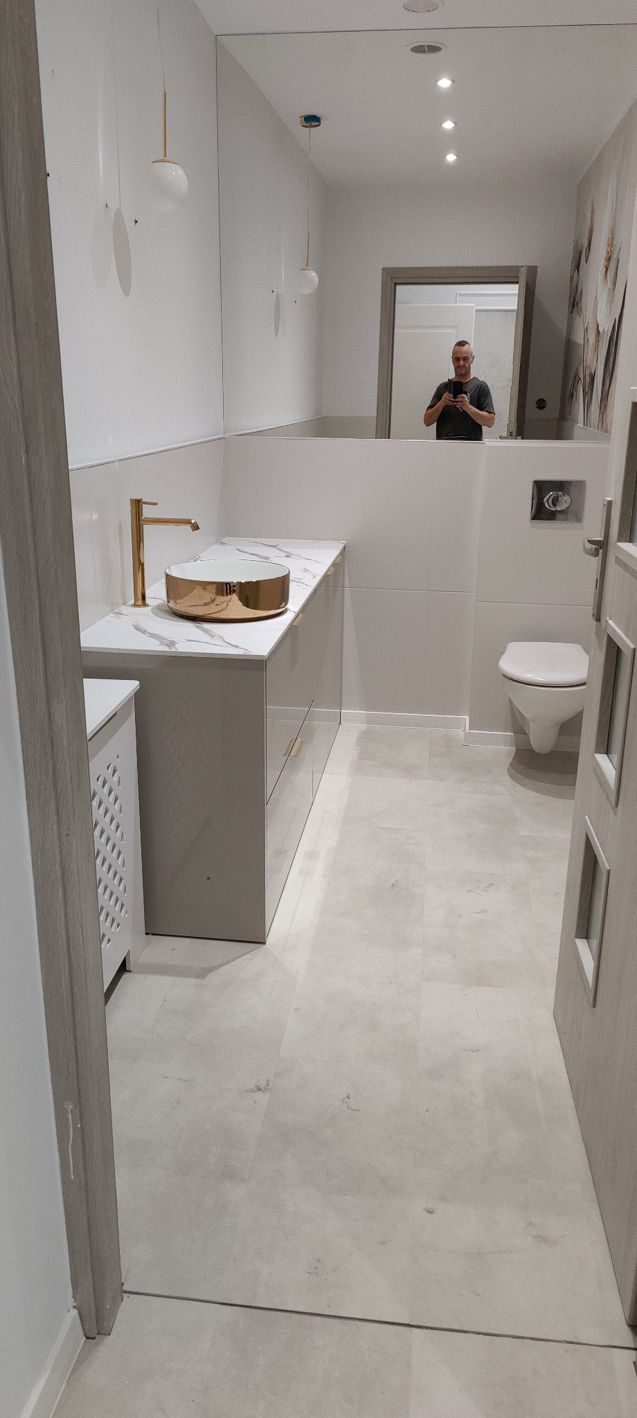 Wykończona łazienka w odcieniach bieli i szarości z elementami złota: umywalka nablatowa w kolorze złotym, armatura w kolorze złotym, duże lustro, toaleta, płytki podłogowe w odcieniach szarości...