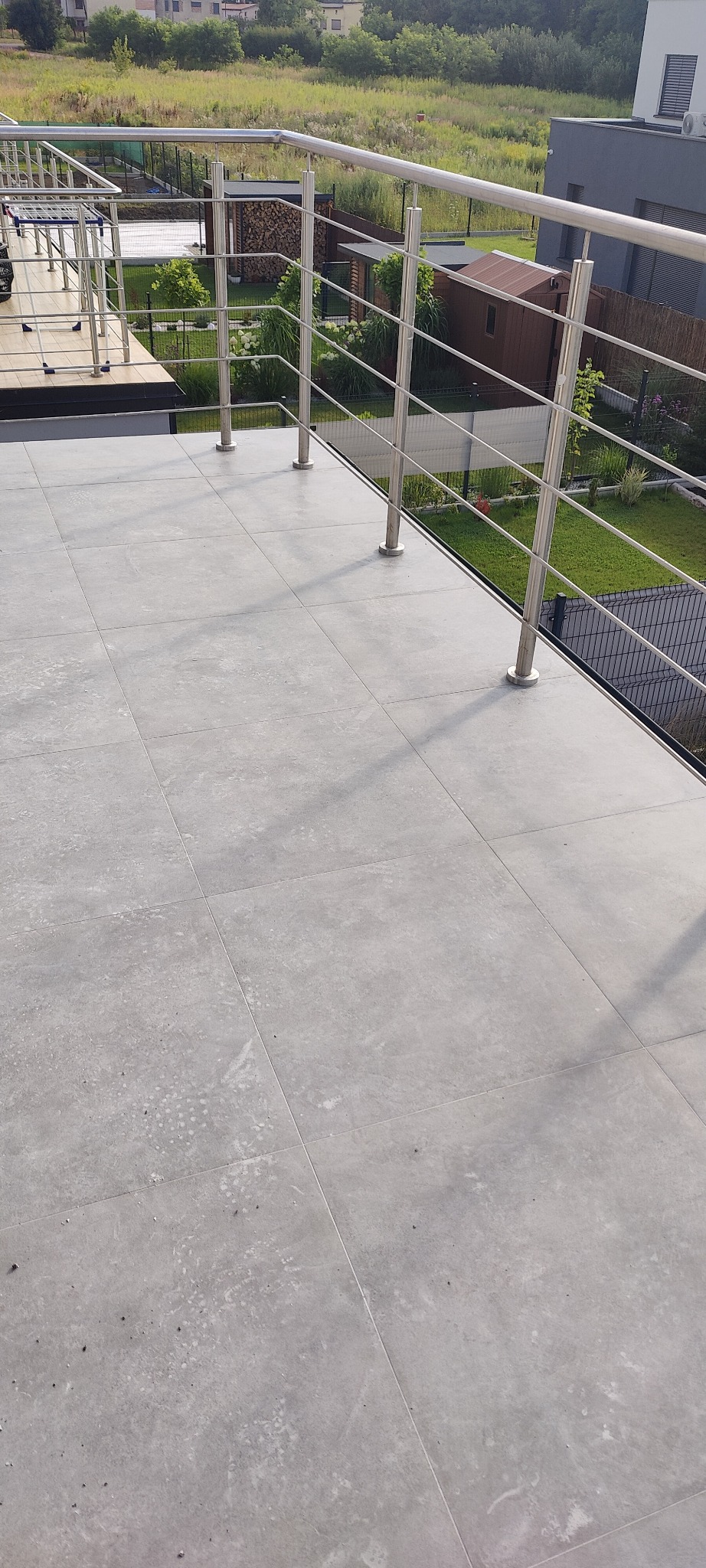 Szare płytki podłogowe na balkonie z metalową balustradą, widok na zielony ogród i zabudowania w tle.