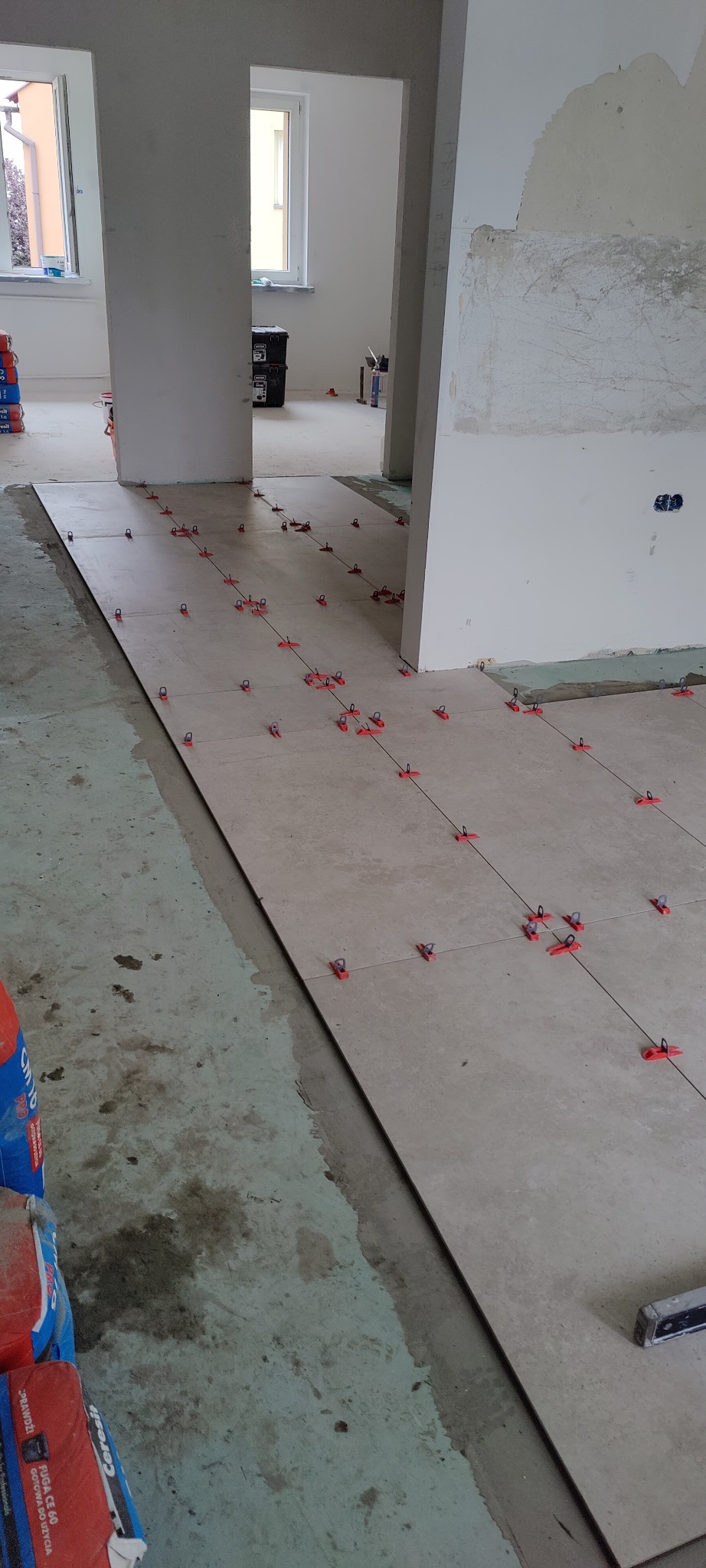 Układanie beżowych płytek podłogowych w trakcie prac wykończeniowych, widoczne krzyżyki dystansowe i worki z zaprawą klejową.