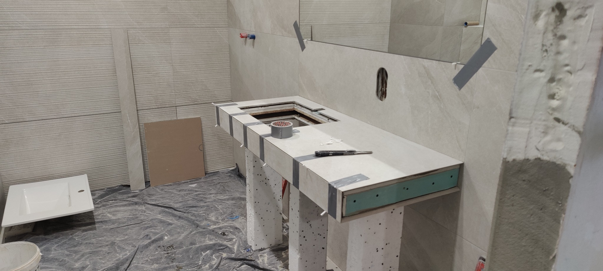 Montaż blatu umywalkowego z płyt kartonowo-gipsowych i płytek w łazience, widoczna taśma maskująca, puszka z klejem i nożyk na blacie, płytki na ścianach z poziomymi paskami, podłoga zabezpieczona...