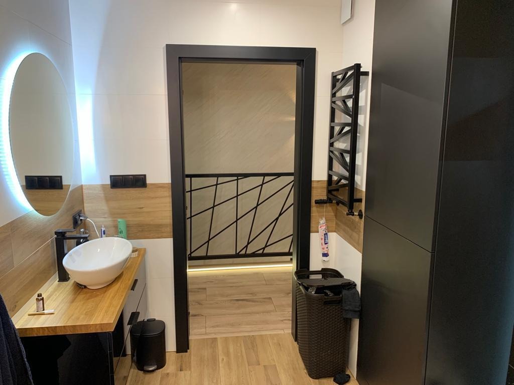 Nowoczesna łazienka z okrągłym lustrem podświetlanym LED, drewnianym blatem, białą umywalką nablatową i czarną armaturą, widoczny grzejnik drabinkowy i kosz na pranie w kolorze ciemnego brązu.