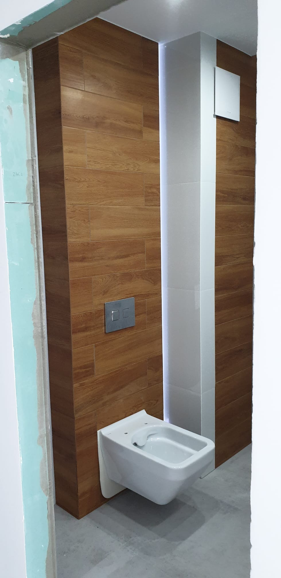 Wnętrze łazienki w trakcie wykańczania, z widoczną podwieszaną miską WC, płytkami imitującymi drewno na ścianie, fragmentem kolumny z białych płytek i surową ścianą z płyty gipsowo-kartonowej.
