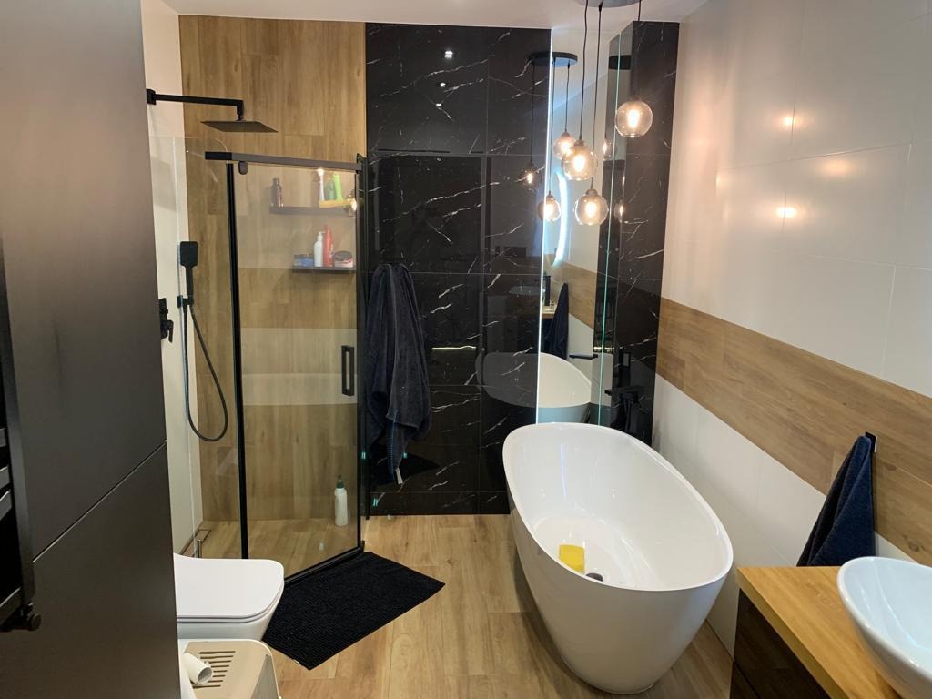 Nowoczesna łazienka z kabiną prysznicową typu walk-in, wolnostojącą wanną i czarno-białą glazurą imitującą marmur, oświetlona wiszącymi lampami z kulistymi kloszami.