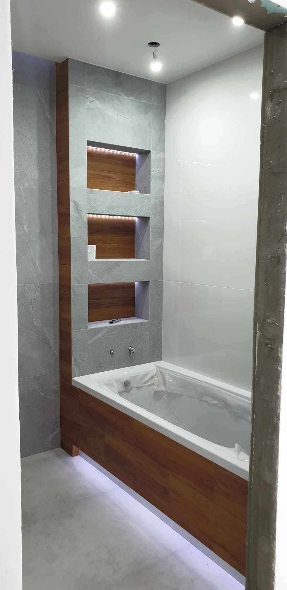 Remont łazienki: zabudowa wanny z drewnopodobną okładziną, szare płytki imitujące kamień z wnękami podświetlanymi LED, białe płytki na ścianie, bateria umywalkowa.