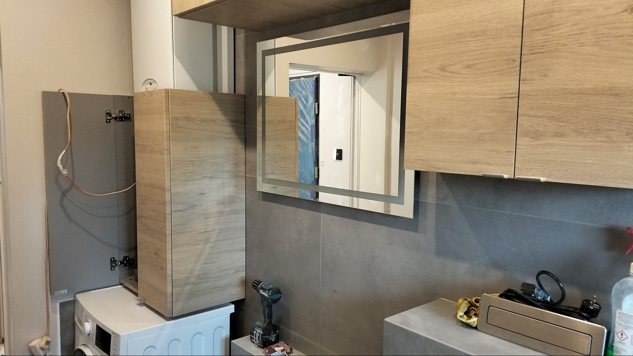 Montaż szafki łazienkowej z lustrem na szarych płytkach, widoczna pralka i wiertarka na blacie.
