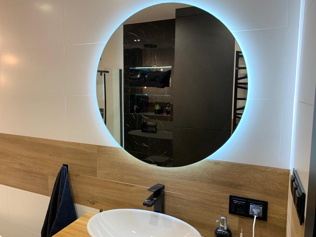 Nowoczesna łazienka z okrągłym lustrem podświetlanym LED, drewnianym blatem pod umywalką i białymi płytkami na ścianach, widoczny czarny kran i dozownik mydła.